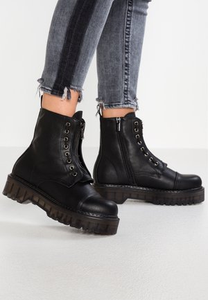 Person iført sorte læderstøvler med snørebånd, lynlåse på siden og kraftige såler, kombineret med grå jeans med frynset kant.