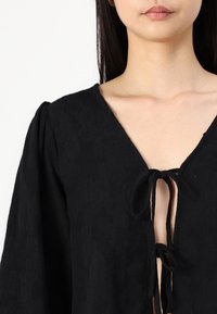 Blouse noire en tissu texturé, col en V profond avec fermetures à nouer à l'avant. Caractérisée par des manches bouffantes et un motif complexe.