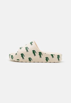 En enkelt beige slip sandal med grønne krokodillemønstre over det hele, vist fra siden på en hvid baggrund.