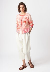 Light pink cardigan with floral patterns in red and orange, round neck, ribbed cuffs, paired with white loose trousers and brown sandals.