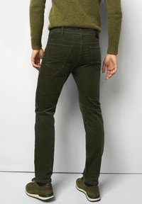 Pantalon en velours côtelé vert avec une coupe slim, comportant des poches arrière et une étiquette à la taille, associé à des chaussures marron. Tissu texturé et design de jambe fuselée.
