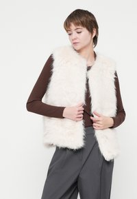 Kamizelka z futra sztucznego w kolorze off-white, z miękką, puszystą teksturą i otwartym przodem, połączona z brązowym, prążkowanym topem z długim rękawem i szarymi spodniami.