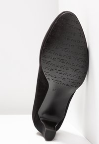 Semelle d'une chaussure à talon haut en daim noir avec "Tamaris" embossé plusieurs fois pour l'adhérence, vue de dessous sur un fond blanc.