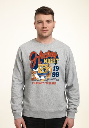 Mężczyzna w szarej bluzie z postacią z kreskówki trzymającą kij hokejowy, tekst "Hockey Night 1999 I'm Ready I'm Ready" i krążki hokejowe.