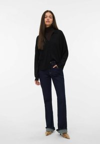 Svart oversized cardigan, mörk polotröja, mörka utsvängda jeans med uppvikta fållar och mönstrade klackar. Släta texturer och subtila färger.