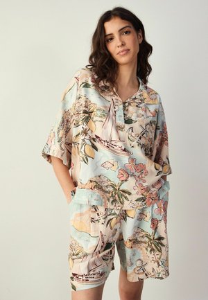 Nő pasztellszínekből álló, rövid ujjú, egyező mintás ingben és rövidnadrágban, hajó-, virág- és trópusi mintával, zsebbe tett kézzel áll.