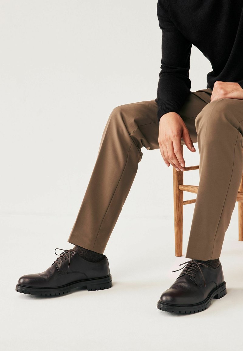 Schwarze Leder-Oxfords mit runden Zehen, Schnürdesign und strukturierter Sohle, getragen mit khakifarbenen Hosen und dunklen Socken, sitzend auf einem Holzstuhl.