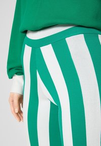 Personne portant un pantalon rayé verticalement vert et blanc et un haut vert à manches longues avec un poignet blanc sur le poignet gauche.