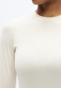 Αθλητική μακρυμάνικη μπλούζα σε ανοιχτό μπεζ χρώμα, κατασκευασμένη από απαλό ύφασμα, με διακριτικό λογότυπο σε λευκό στο στήθος και επίπεδες ραφές για άνεση.