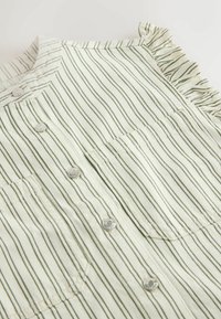 Tissu blanc à fines rayures verticales vertes, boutons-pression argentés sur le devant, poche carrée et détail froncé à l'épaule sur un vêtement.