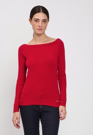 Kvinde med mørkt hår i lav hestehale iført en tætsiddende rød sweater med bare skuldre og mørkeblå jeans, stående foran en ensfarvet hvid baggrund.