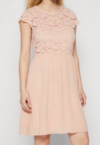 Robe rose clair avec un corsage en dentelle à manches courtes et une jupe fluide et transparente. Le tissu a une texture lisse avec des motifs floraux.