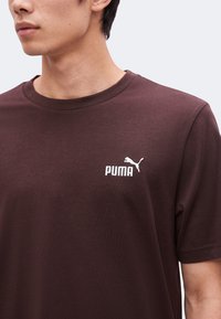Νέος άνδρας που φοράει ένα απλό σκούρο καφέ T-shirt Puma με λευκό λογότυπο Puma στο αριστερό μέρος του στήθους, με λευκό φόντο.