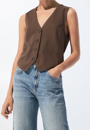 Personne portant un gilet marron sans manches à boutons et un jean taille haute bleu clair, une main dans la poche, sur fond uni.