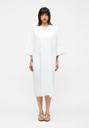 Žena stojaca tvárou vpredu v širokých bielych midi šatách so širokými rukávmi a hnedými sandálmi na remienky na jednofarebnom bielom pozadí.