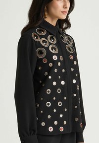 Maglietta nera con paillettes circolari in argento, nero e rosso, chiusura con bottoni e maniche lunghe. Superficie testurizzata con decorazioni.