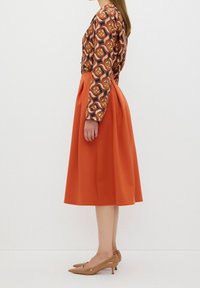 Blus med långa ärmar i brunt och orange geometriskt mönster, tillsammans med en orange A-linje kjol. Modellen bär ljusbruna spetsiga klackar.