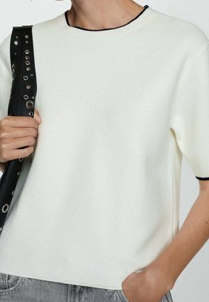 Person trägt eine weiße kurzärmelige, strukturierte Oberteil mit schwarzem Rand und hält eine schwarze Tasche mit silbernen Ösen über einer Schulter.