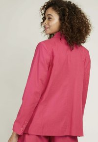Blazer rose ajusté avec un col structuré, des manches longues et des accents de boutons sur les poignets. Tissu lisse au design épuré et minimal.