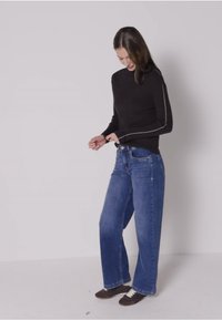 Zwarte langemouwen shirt met witte stiksels, gecombineerd met brede blauwe jeans. Model staat in een neutrale omgeving en toont casual kleding en schoeisel.