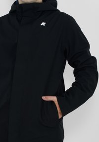 Veste à capuche noire au design élégant. Elle présente une fermeture éclair, des poches latérales et un petit logo sur la poitrine. Texture douce et mate.