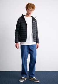 Zwarte pufferjack met horizontale stiksels en capuchon, gecombineerd met een wit T-shirt en wijde denim jeans. Gestyled met grijze sneakers.