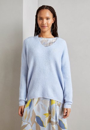 Hellblauer, locker sitzender Pullover aus weichem, strukturiertem Stoff mit tiefem V-Ausschnitt. Getragen über einem gemusterten Rock mit blauen, gelben und beigefarbenen Akzenten.