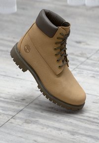 Καστόρινη δερμάτινη μπότες με κορδόνια, επενδυμένη με σκούρο καφέ μαξιλαράκι στον γιακά και χοντρή λαστιχένια σόλα, που ξεκουράζεται σε ανοιχτό γκρι δαπέδο πλακιδίων.