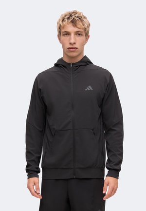 Young man wearing a black zip-up hoodie with a hood and pockets, standing against a plain light background, facing forward.