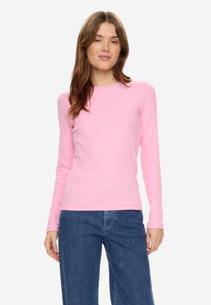 Vrouw met lang bruin haar die een roze shirt met lange mouwen en een blauwe spijkerbroek draagt, staand tegen een effen witte achtergrond.