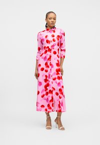 Γυναίκα που φοράει ροζ φόρεμα με κόκκινα και ροζ μοτίβα από πέταλα, μανίκια τρία τέταρτα, κουμπωτό μπροστά, και λευκά σανδάλια με ψηλό τακούνι, στέκεται σε λευκό φόντο.