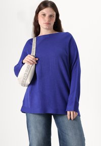 Blauer gestrickter oversized Pullover mit strukturierten Mustern, weiter Ausschnitt und langen Ärmeln, kombiniert mit hellen Jeans und einer kleinen weißen Tasche.