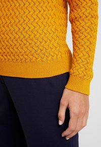 Gul stickad tröja med ett strukturerat diamantmönster, ribbade ärmslut och nederkant, i kombination med mörk marinblå tyg, som visar detaljer av arm och hand.