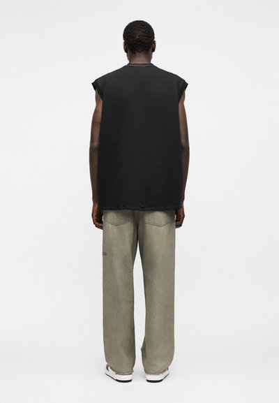 Άτομο όρθιο με την πλάτη γυρισμένη, φοράει μαύρο αμάνικο μπλουζάκι, φαρδιά γκρι παντελόνια και λευκά αθλητικά παπούτσια σε απλό λευκό φόντο.