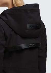 Sort vandtæt jakke med hætte, der har et tekstureret logo-bånd og glat stof. Designet er strømlinet med minimalistiske accenter.