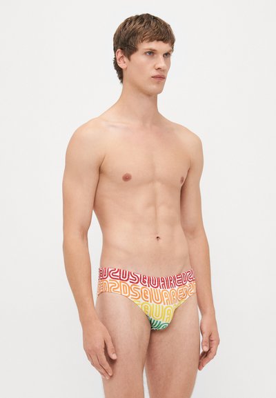 Junger Mann mit kurzen braunen Haaren, der farbenfrohe Schwimmslips mit Dsquared2-Logo-Muster trägt, steht vor einem schlichten weißen Hintergrund.