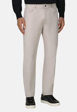 Uomo che indossa pantaloni beige a gamba dritta con tasche frontali, maglione nero e scarpe da ginnastica nere con suola bianca, in piedi davanti a uno sfondo semplice.