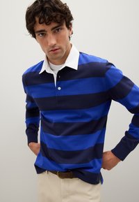 Junger Mann mit lockigem Haar, der ein blaues und navy gestreiftes Langarmpolo mit einem weißen Kragen und beigen Hosen trägt und in die Kamera schaut.