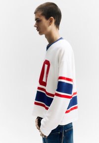 Fehér hosszú ujjú ing piros "0" számmal, piros és kék csíkokkal. Kék farmerral, melyen láncos díszítés található. Oldalnézeti kép.