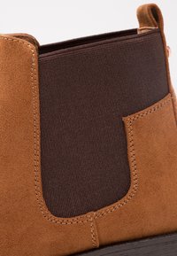 Botín de ante marrón hasta el tobillo con paneles laterales elásticos y detalles de costura. Textura suave con un tacón ligeramente elevado y puntera redondeada.