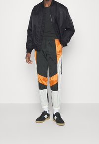 Giacca bomber in satin nera sopra una maglietta nera. I pantaloni presentano pannelli arancioni e bianchi su materiale nero. Sneakers nere con suole in gomma.