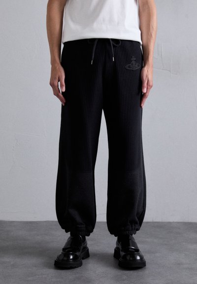 Pantalons de jogging noirs côtelés avec des poignets élastiques, une taille à cordon de serrage et un logo discret sur la cuisse, associés à des chaussures noires brillantes.