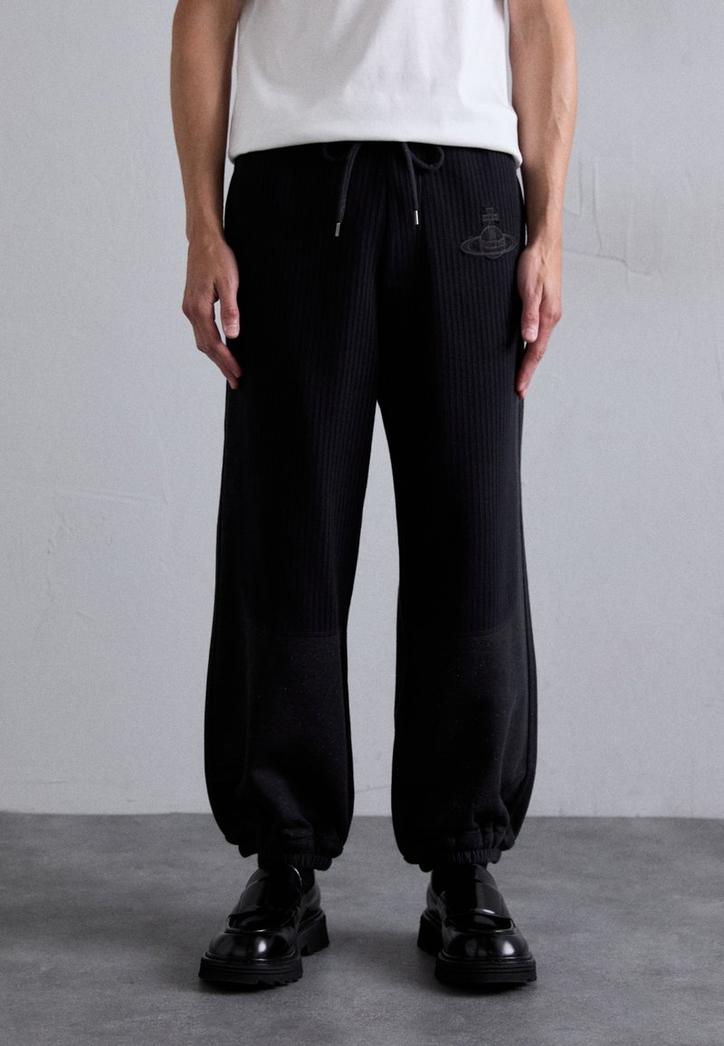 Pantalons de jogging noirs côtelés avec des poignets élastiques, une taille à cordon de serrage et un logo discret sur la cuisse, associés à des chaussures noires brillantes.
