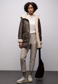 Frau mit lockigem Haar, die eine braune Schaffelljacke, einen weißen Rollkragenpullover, beige Hose, Turnschuhe trägt und eine dunkelbraune Umhängetasche hält.