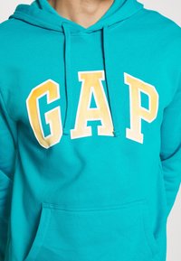 Sudadera con capucha turquesa con bolsillo frontal tipo canguro, capucha ajustable con cordón y un gran gráfico amarillo de "GAP" en letras llamativas.