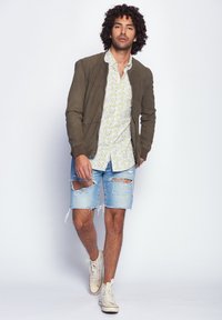 Braune Bomberjacke, weißes Blumenshirt, ausgebleichte hellblaue Shorts und beigefarbene Sneakers mit braunen Akzenten. Model läuft nach vorne.