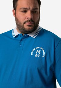 Modrá polo košile z bavlny, s bílým límečkem se dvěma pruhy, knoflíky a logem "Mountain Moments" s číslem 97.
