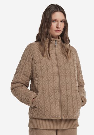 Kvinde iført en beige kabelstrikket lynlåstrøje med høj krave og lommer, kombineret med matchende ribstrikkede bukser.