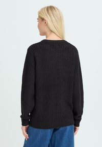 Suéter de punto negro con superficie texturizada, corte relajado, escote redondo y puños y dobladillo de canalé, combinado con pantalones de mezclilla azul.