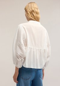 Bluză albă cu mâneci texturate cu dungi, spate strâns și tiv rotunjit. Asortată cu blugi albaștri denim, evidențiind o croială lejeră.